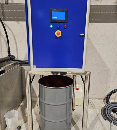 Panel de control industrial de color azul con una pantalla digital encendida, un botón de parada de emergencia rojo y luces indicadoras. El armario está montado sobre una estructura de acero inoxidable con un bidón metálico gris situado debajo de él.